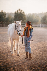 A young cowgirl woman stroking a horse in the sunset at her ranch.