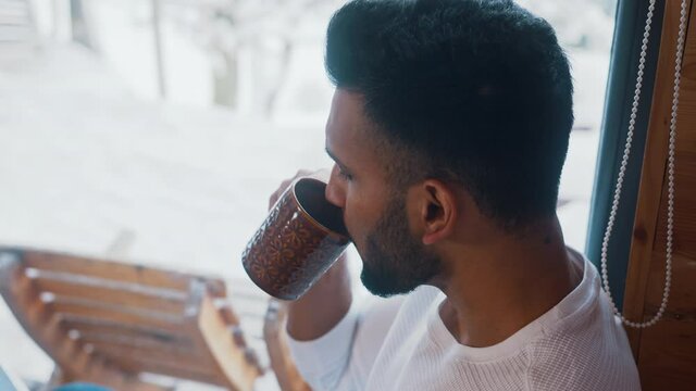 Close up. Young indian man drinking coffee near the window and looking at snow outside. High quality 4k footage