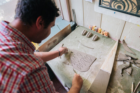 Young man working with clay and creating small ceramic objects in home studio workshop