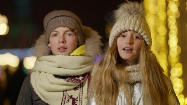 Happy Caucasian boy and girl singing Christmas carol with golden New Year lights at background in decorated city outdoors. Cheerful children enjoying holiday celebration. Childhood and lifestyle.