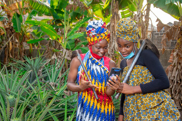 Two beautiful African female farmers with nose mask, looking happily into a smartphone and holding hoe and cutlass in a banana plantation  