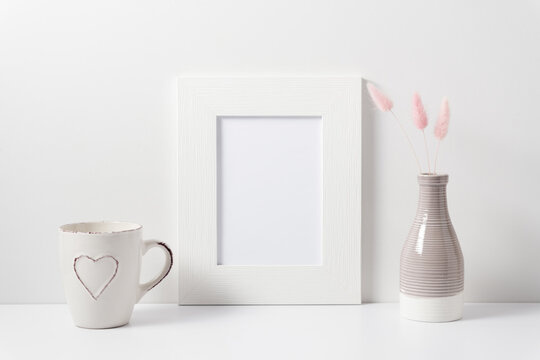 White blank photo frame, dry flowers in vase, cup on white table. Front view. Place for text, copy space, mockup