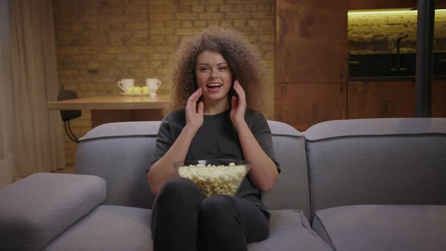 Pretty curly haired woman laughing in front of tv at home. Millennial woman watching comedy movie and eating pop corn sitting on couch.