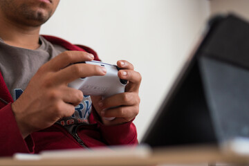 Young man wearing a red jacket playing videogames with a white controller on his black tablet at home 