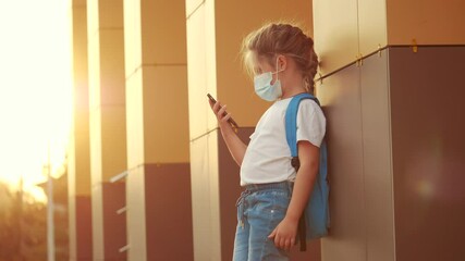 schoolgirl in a protective mask. child with a backpack stands near the school with a smartphone. schoolgirl education in a protective mask outdoors. coronavirus pandemic covid 19 concept
