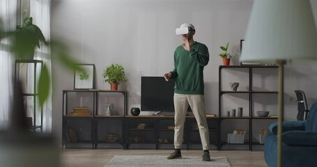young man is using head-mounted display standing in living room and swiping virtual screen by hands, full-length