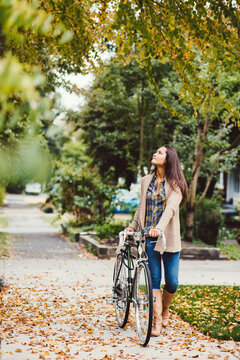 Young woman walking down leaf covered sidewalk with bike.