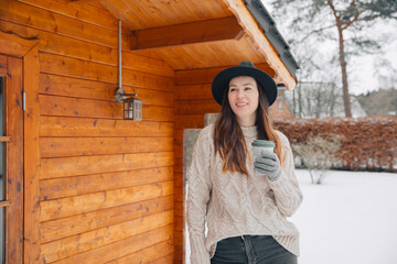 woman in the snow. Woman in a knitted sweater and black hat holds a cup of tea in hand. Woman outside in winter. Vacation in a wooden house in winter. Enjoy life in winter	
