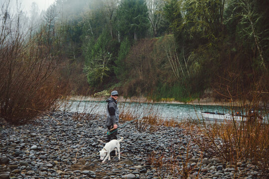 Fly fisherman walking along the rocky shore of a river with his dog