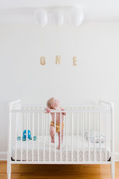 Cute baby standing in a crib on her first birthday with balloons and the word one
