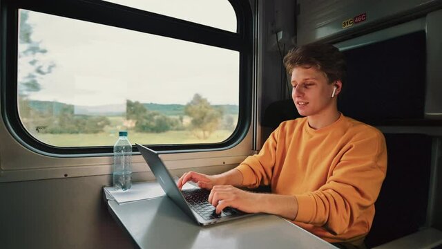 Handsome young man rides a train and works at a desk on a laptop with a smile on his face communicating on wireless headphones.