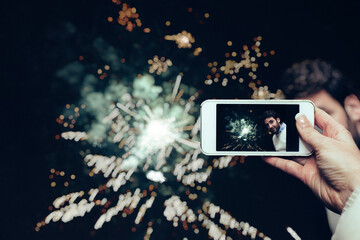 A female hand holding a mobile phone, and taking a photo of a man in front of fireworks