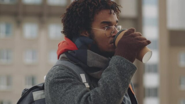 Side view of young black man in outerwear standing outdoors in city, drinking coffee from to go cup and looking away on cold winter day