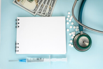 A few hundred dollar bills pills and a stethoscope on a blue background near the notebook. Space for the text.Medical concept