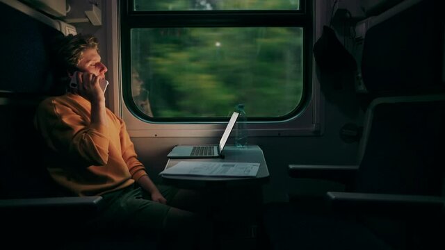Positive young man sitting with a laptop at a table in a train compartment and calling on the phone while working online on a trip.