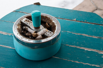 A retro, antique cigarette ashtray filled with stubs on a turquoise sanded wooden table top. 