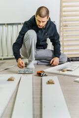 One Man with beard alone Putting Together Self Assembly Furniture sitting on the floor at Home holding electric screwdriver looking the instructions - side view DIY concept real people copy space