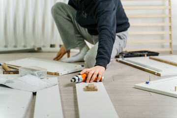 One unknown Man alone Putting Together Self Assembly Furniture sitting on the floor at Home holding electric screwdriver looking the instructions - side view DIY concept real people copy space