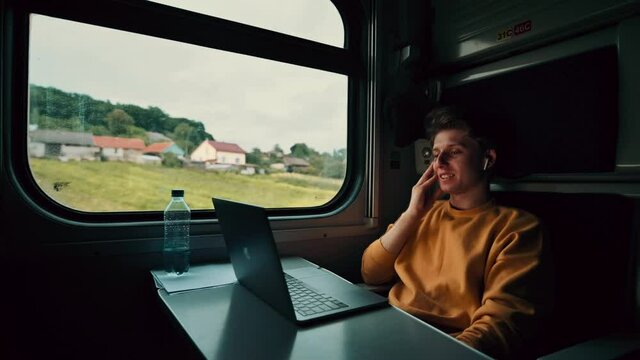 Happy guy in wireless headphones watching funny video on laptop in train while traveling. Tourist on a train watches a comedy movie and laughs.