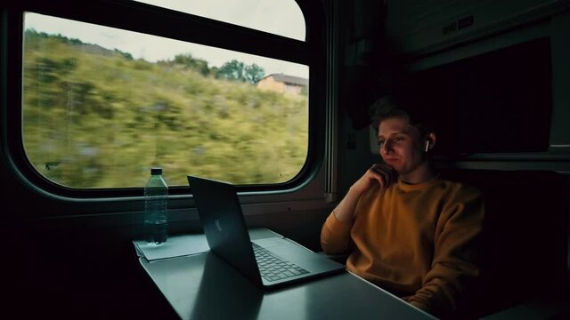 Positive young man sitting in the evening compartment of a train and watching a movie on a laptop with a smile on his face. Guy travels and watches content on a laptop.