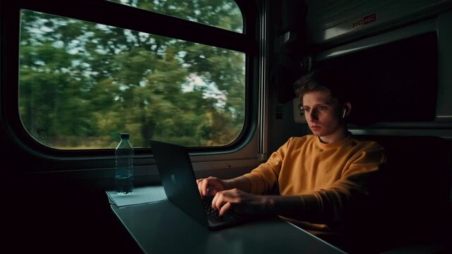 Concentrated young man in wireless headphones sitting at a table in a train compartment and working on a laptop.