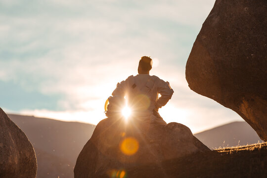 A weary astronaut rests on a rock looking out on the distance of a foreign horizon