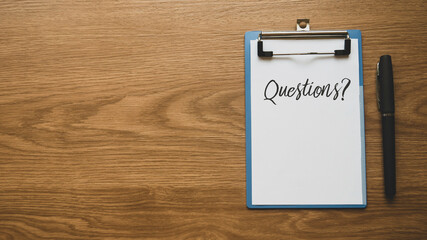 Top view of a pen and clipboard with paper written with Questions and symbol on wooden background...