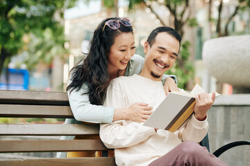 Pretty smiling young Asian woman looking in book of boyfriend reading book when sitting on park...
