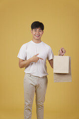 portrait of happy young asian man dressed casually holding shopping bags isolated on yellow background