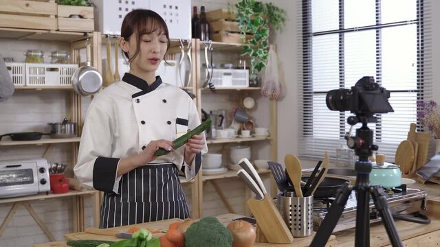 beautiful asian culinary specialist vlogging at home. introducing ingredients with gestures before cooking a meal, holding cucumber and onion in front of digital camera.