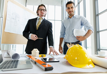 Young engineer standing together with older boss in a suit at office with roll of plan paper and toll for engineer related field..