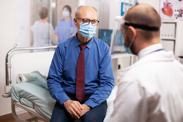 Obraz premium Doctor in the course of covid outbreak consulting senior man in examination room. Physician practitioner in the course of private modern clinic appointment during coronavirus pandemic.