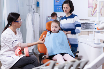 Obraz premium Child wearing dental bib sitting on chair while doctor is showing artificial teeth. Little girl and mother listening stomatolog talking about tooth hygine in dentistiry clinic holding jaw model.