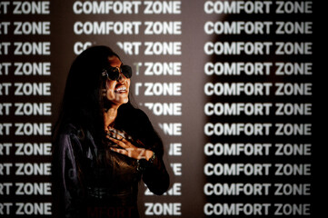 Young indian model with sunglasses and black jacket looking at camera. Woman smiling in dark studio illuminated by light of projector with word comfort zone