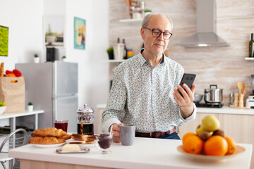 Happy old man surfing on social media using smartphone during breakfast sitting in kitchen smiling....