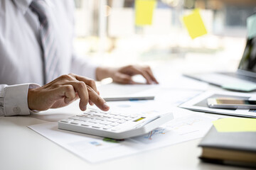 A businessman is using a white calculator to calculate financial numbers, he is sitting in his...