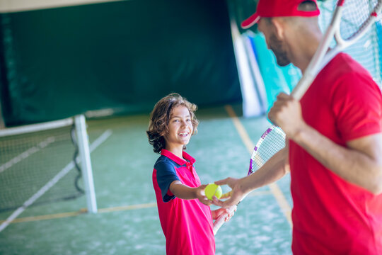 Young bearded man in a red cap shaking hands with a dark-haired boy before playing tennis