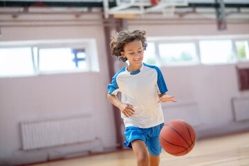 Dark-haired boy in sportswear running after the ball in a gym and looking concentrated