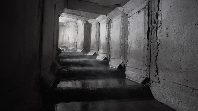 Square concrete drainage underground tunnel with steam evaporating from the water. Concrete sewer tunnel turning with steps, with the light shines behind the turning.