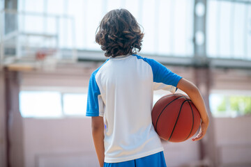 Dark-haired boy in sportswear holding a ball and looking at the basketball ring