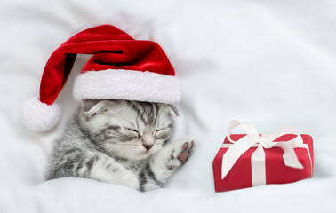 Tabby kitten wearing red santa's hat sleeps with gift box under a white blanket on a bed. Top down view. Empty space for text