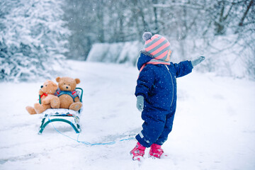 Full growth photo of toddler girl in a blue overalls, ultimate gray mittens, scarf, hat, shoes stands near a sleigh with teddy bears, winter outdoors. Kid smiling with tongue out, showing in distance