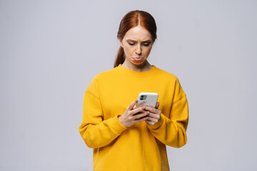 Displeased cute young woman reading bad news, feels dissatisfaction after receiving message on isolated white background. Pretty lady model with red hair emotionally showing facial expressions