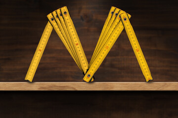 Yellow old wooden folding ruler in the shape of letter M. Meter, unit of length. Above a wooden shelf. Photography.