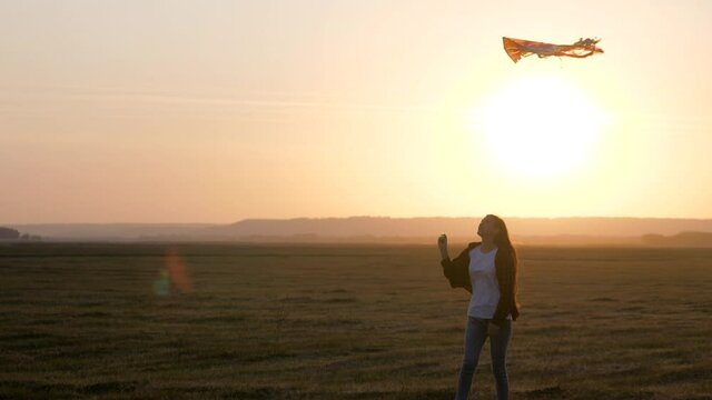 Girl plays with multi-colored kite on field in rays of sun. Teenager dreams of flying and becoming pilot. colored kite hangs in air. Kite Festival. Child and flying kite at sunset in summer.