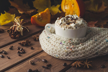 Cup of coffee with cinnamon sticks, whipped cream and chocolate sprinkles. Mug covered with knitted winter scarf. Cozy autumn composition of hot beverage on wooden table.