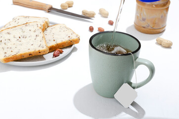 Closeup of pouring hot water into the cup, process of making tea and plate with fresh slices of bread, jar of peanut butter on the white table