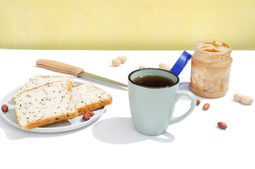 Breakfast time.Cup of black tea, fresh slices of bread, peanut butter, peanuts, knife on the white table against yellow background