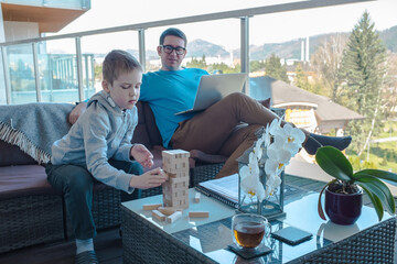 Stay and work at home dad. Father and son spend free time on terrace. Man working, child boy playing, builds, creating, wooden game toy. Weekend Family. Bonding concept.
