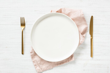 White empty plate and golden cutlery with pink napkin on white wooden table. Top view on table setting.
